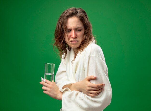 Weak young ill girl looking down wearing white robe holding glass of water crossing hands isolated on green