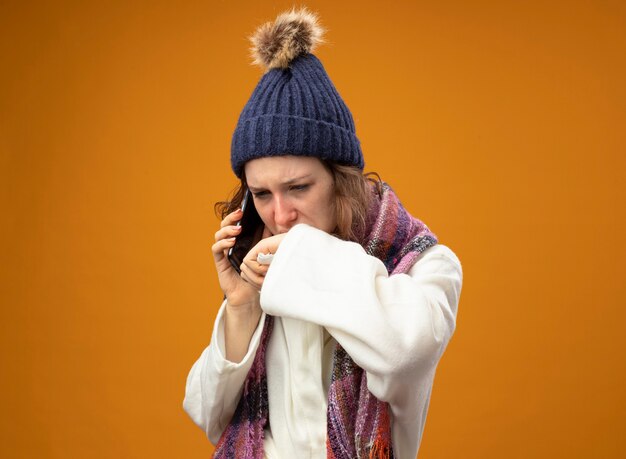 weak young ill girl looking at down wearing white robe and winter hat with scarf speaks on phone wiping mouth with hand isolated on orange
