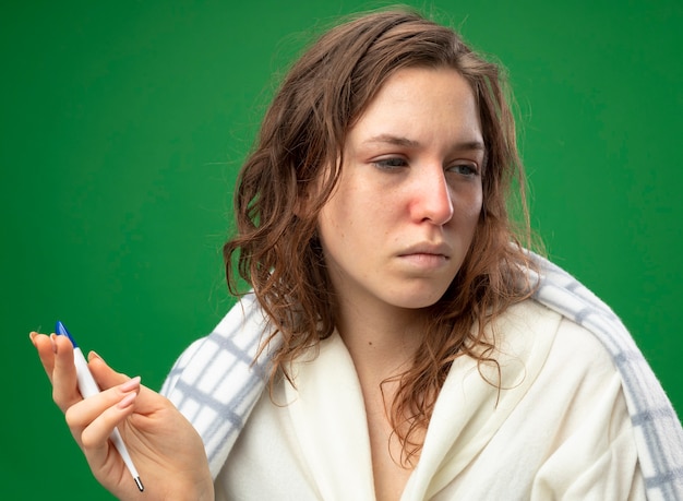 Слабая молодая больная девушка смотрит в сторону в белом халате, завернутом в плед, с термометром, изолированным на зеленом