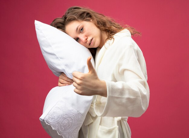 Weak young ill girl looking straight ahead wearing white robe hugged pillow showing thumb up isolated on pink