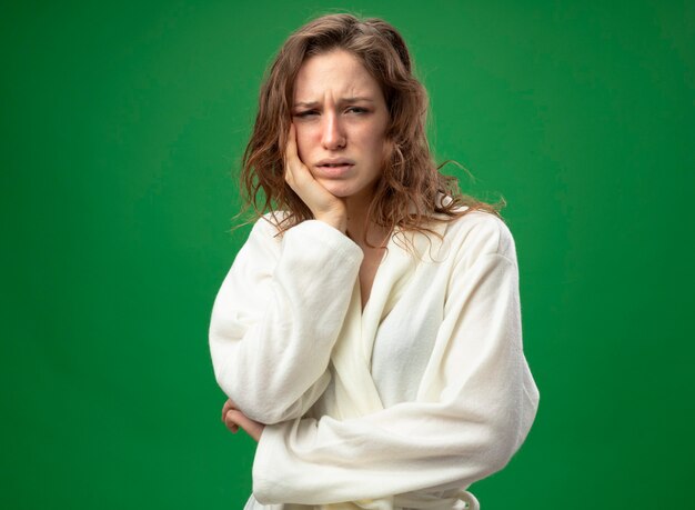 Weak young ill girl looking straight ahead wearing white robe putting hand on cheek isolated on green