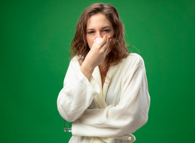 Weak young ill girl looking straight ahead wearing white robe wiping nose with napkin isolated on green