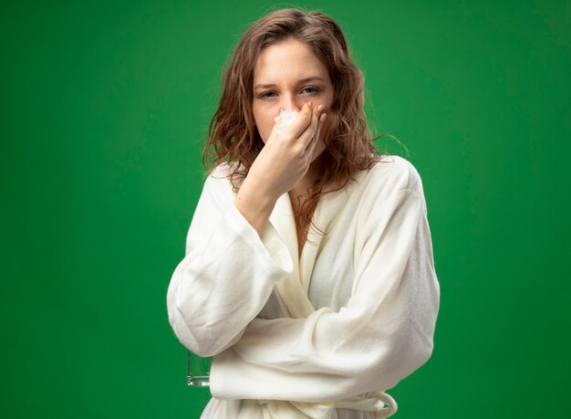 Weak young ill girl looking straight ahead wearing white robe wiping nose with napkin isolated on green