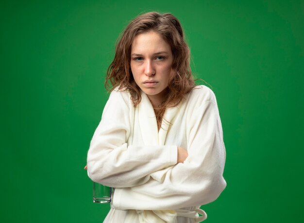 Weak young ill girl wearing white robe holding glass of water crossing hands freezing cold isolated on green