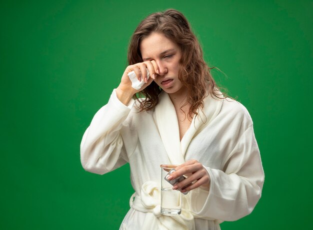 Слабая молодая больная девушка в белом халате, держащая стакан воды с таблетками и вытирающая глаза рукой, изолированной на зеленом с копией пространства