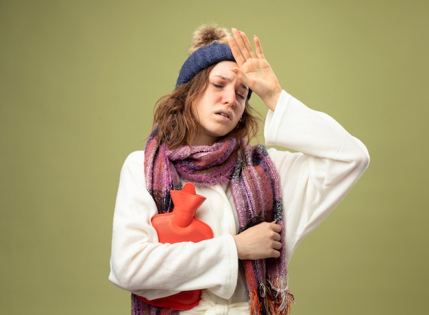 Foto gratuita debole giovane ragazza malata che indossa una veste bianca e cappello invernale con la sciarpa che tiene la borsa dell'acqua calda che mette la mano sulla fronte isolata su verde oliva