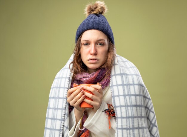 Weak young ill girl wearing white robe and winter hat with scarf wrapped in plaid holding cup of tea isolated on olive green