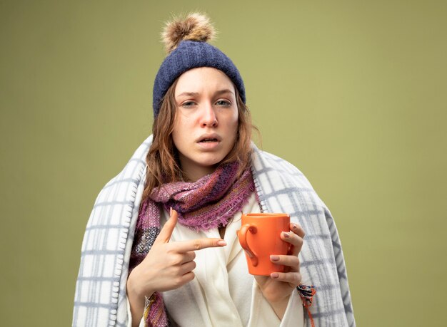 Слабая молодая больная девушка в белом халате и зимней шапке с шарфом, завернутым в клетчатый холдинг, и указывает на чашку чая, изолированную на оливково-зеленом