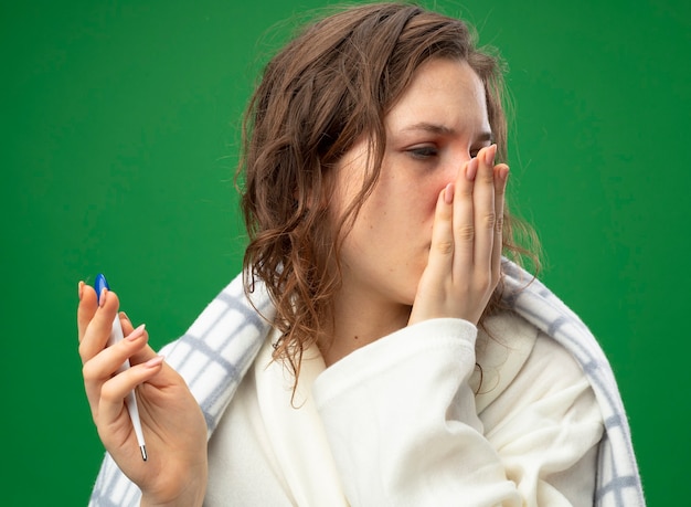 Weak young ill girl wearing white robe wrapped in plaid holding thermometer covered mouth with hand isolated on green