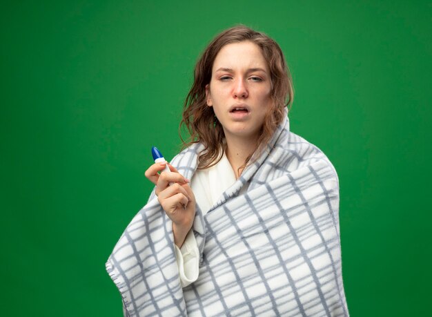 Слабая молодая больная девушка в белом халате, завернутая в плед, с термометром, изолированным на зеленом