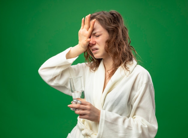 Weak young ill girl with closed eyes wearing white robe holding glass of water putting hand on forehead isolated on green