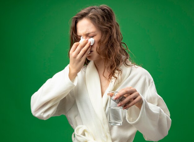 緑に分離されたナプキンで水拭き鼻のガラスを保持している白いローブを着て目を閉じて弱い若い病気の女の子