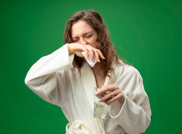 目を閉じて弱い若い病気の女の子は、丸薬と水のガラスを保持し、緑に分離された手で鼻を拭く白いローブを着ています