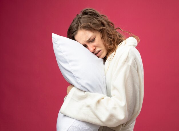 Weak young ill girl with closed eyes wearing white robe hugged pillow isolated on pink