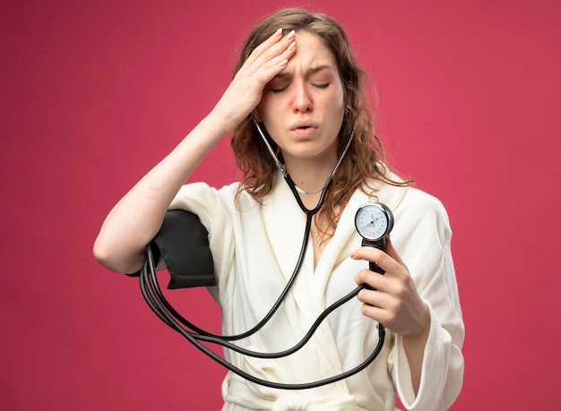 Free photo weak young ill girl with closed eyes wearing white robe measuring her own pressure with sphygmomanometer putting hand on forehead isolated on pink