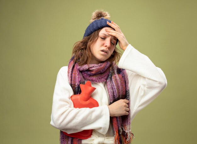 weak young ill girl with closed eyes wearing white robe and winter hat with scarf holding hot water bag putting hand on forehead isolated on olive green
