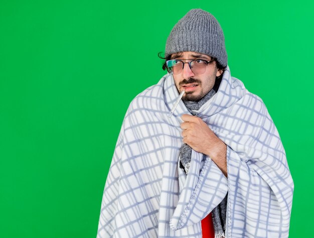 Weak young ill man wearing glasses winter hat and scarf wrapped in plaid holding thermometer in mouth looking at side grabbing plaid isolated on green wall