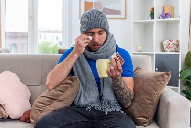 weak young ill man wearing scarf and winter hat sitting on sofa in living room holding packs of pills and cup of tea wiping eye with closed eyes