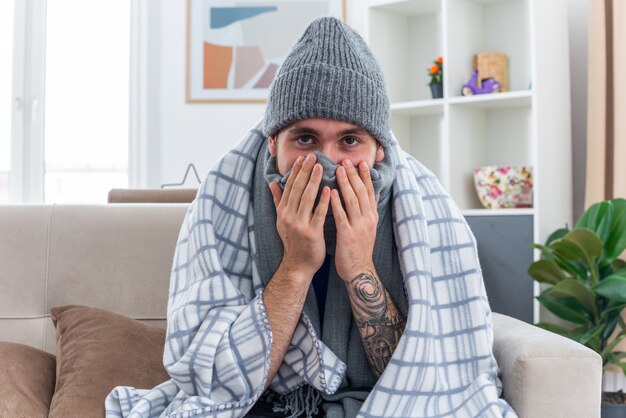weak young ill man wearing scarf and winter hat sitting on sofa in living room wrapped in blanket covering mouth and nose with scarf keeping hands on face looking at front