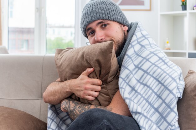 Weak young ill man wearing scarf and winter hat sitting on sofa in living room wrapped in blanket hugging pillow looking at camera