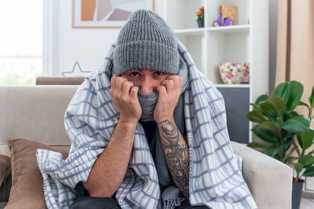 Weak young ill man wearing scarf and winter hat sitting on sofa in living room wrapped in blanket looking at camera covering mouth with scarf