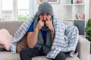 Free photo weak young ill man wearing scarf and winter hat wrapped in blanket sitting on sofa in living room looking at front pointing fingers on nose