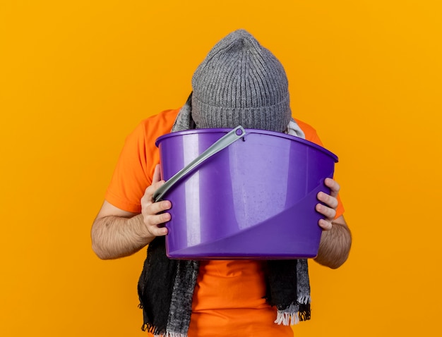 Слабый молодой больной в зимней шапке с шарфом, держащий пластиковое ведро и рвущий в него, изолированный на оранжевом
