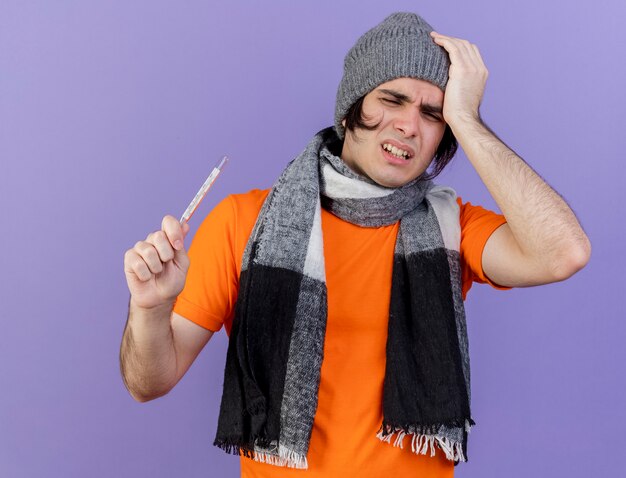 weak young ill man wearing winter hat with scarf holding thermometer and putting hand on aching head isolated on purple