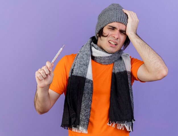слабый молодой больной мужчина в зимней шапке с шарфом держит термометр и кладет руку на больную голову, изолированную на фиолетовом