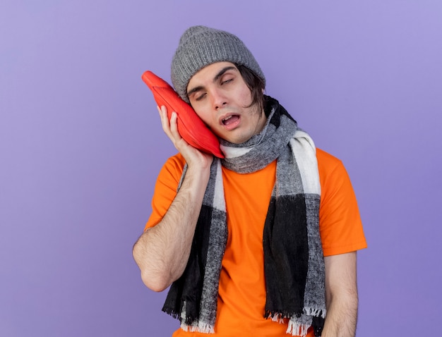 weak young ill man wearing winter hat with scarf putting hot water bag on cheek isolated on purple