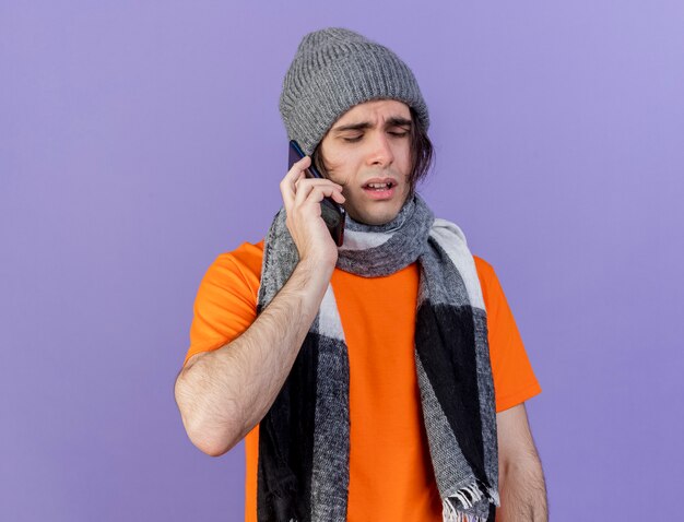 weak young ill man wearing winter hat with scarf speaks on phone isolated on purple