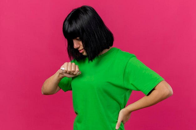 Weak young ill woman standing in profile view keeping hand on waist holding napkin looking down isolated on pink wall with copy space