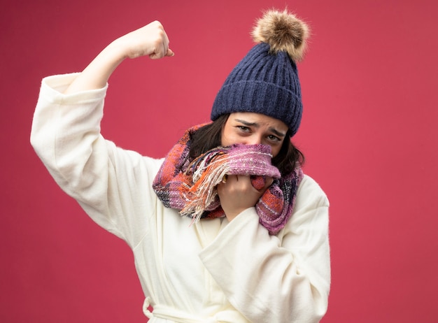 Слабая молодая больная женщина в зимней шапке и шарфе, закрывающем рот шарфом, смотрит вперед, делая сильный жест, изолированный на розовой стене