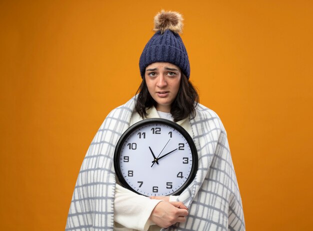 Weak young ill woman wearing robe winter hat wrapped in plaid holding clock looking at front isolated on orange wall