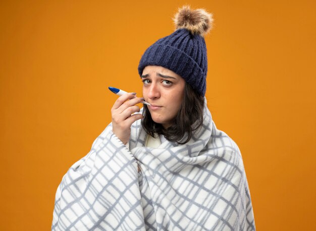 Weak young ill woman wearing robe winter hat wrapped in plaid putting thermometer in mouth looking at front isolated on orange wall