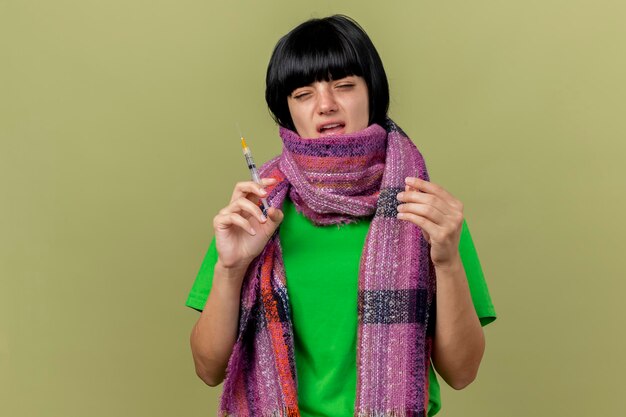 Weak young ill woman wearing scarf holding syringe and ampoule looking at front isolated on olive green wall with copy space