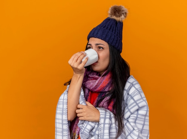 縦断ビューで立っている格子縞に包まれた冬の帽子とスカーフを身に着けている弱い若い病気の女性オレンジ色の壁に孤立してまっすぐに見えるお茶を飲む