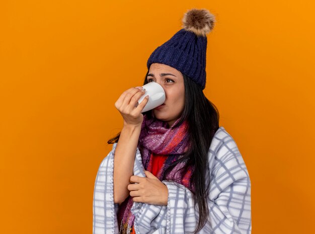 縦断ビューで立っている格子縞に包まれた冬の帽子とスカーフを身に着けている弱い若い病気の女性オレンジ色の壁に孤立してまっすぐに見えるお茶を飲む