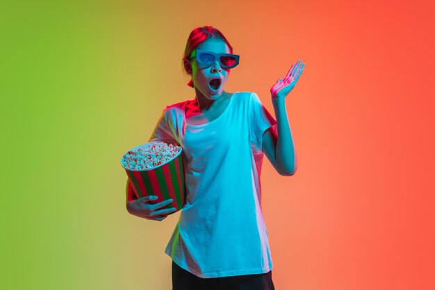 Wearing 3Dheadset shocked scared with popcorn Young caucasian girls portrait on gradient greenorange studio background in neon light Concept of youth human emotions facial expression ad