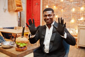 食べ物のための手袋を着用しておいしいダブルハンバーガーとレストランに座っている黒いスーツを着た立派な若いアフリカ系アメリカ人の男