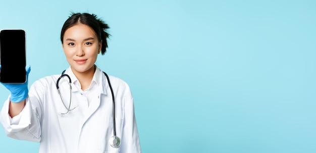 Бесплатное фото Концепция веб-врача улыбающийся азиатский врач показывает экран мобильного телефона, стоящий в клинике оон