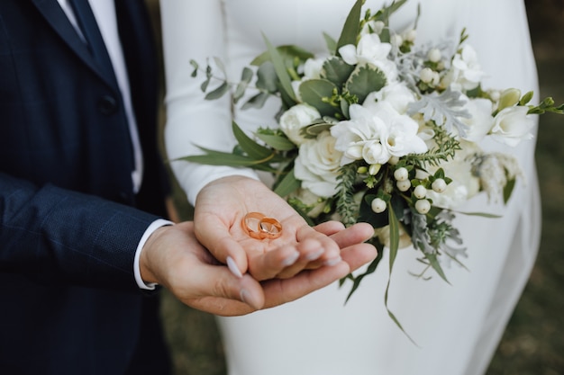 Wedding bands in the hands of bride and groom and with beautiful wedding bouquet made of greenery and white flowers