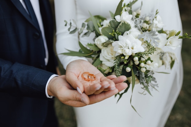 신부와 신랑의 손에 녹지와 흰색 꽃으로 만든 아름다운 웨딩 부케와 웨딩 밴드