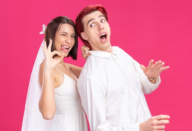 Wedding couple groom and bride in white wedding dress , bride holding collar of her confused groom , doing ok sign happy and cheerful