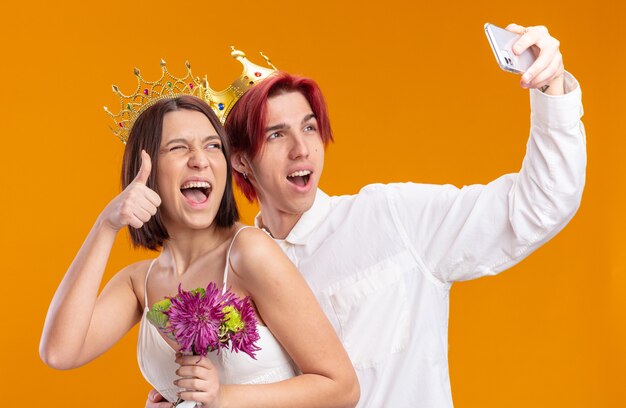 Wedding couple groom and bride with bouquet of flowers in wedding dress wearing gold crowns smiling cheerfully doing selfie using smartphone showing thumbs up