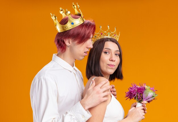 Wedding couple groom and bride with bouquet of flowers in wedding dress wearing gold crowns smiling cheerfully posing together happy in love