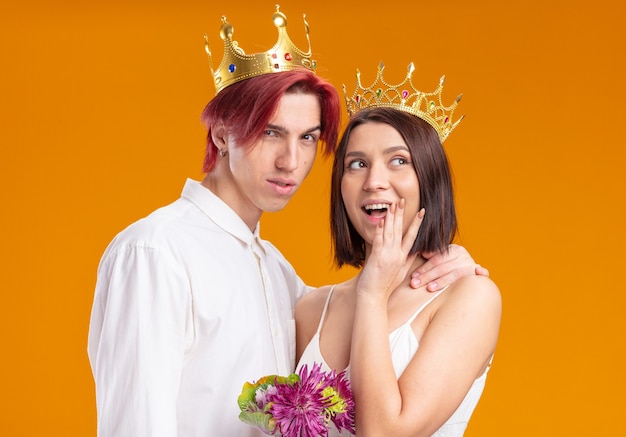 一緒に元気にポーズをとって笑顔の金の王冠を身に着けているウェディングドレスの花の花束を持つ結婚式のカップルの新郎と新婦