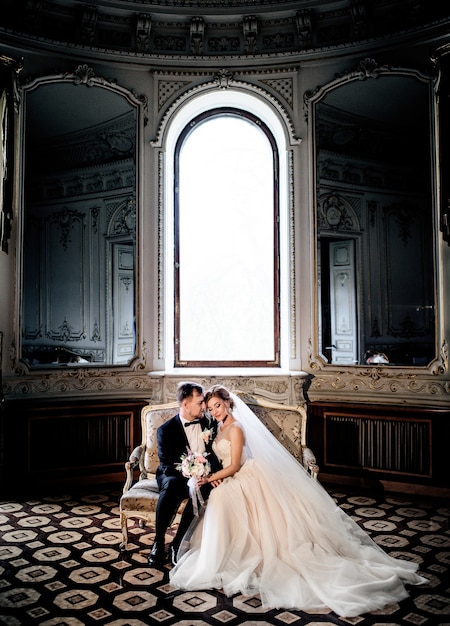 wedding couple hugs on the couch sitting before a tall bright window in a luxury old hall