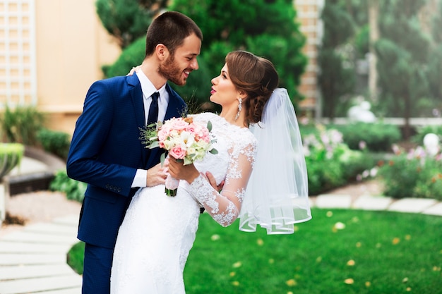 愛のキスと笑顔の結婚式のカップル。若いかなりエレガントな花嫁と緑豊かな公園でポーズをとる彼女のハンサムな新郎。