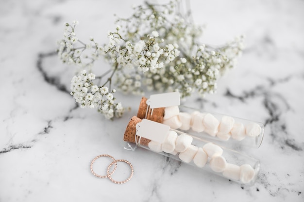 Wedding rings; marshmallow test tubes with tag and baby's-breath flowers on white textured background